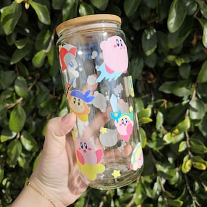 May include: A clear glass water bottle with a bamboo lid and colorful cartoon character stickers. The stickers feature pink, yellow, blue, and white characters with stars and clouds.