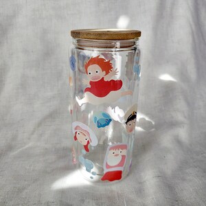 K&ouml;nnte beinhalten: Ein klares Glas mit Bambusdeckel, verziert mit Aufklebern von Zeichentrickfiguren. Die Aufkleber zeigen eine rot gekleidete Figur in der Luft, zusammen mit anderen animierten Figuren. Der zylindrische Becher ist leer.