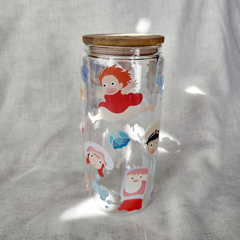 K&ouml;nnte beinhalten: Ein klares Glas mit Bambusdeckel, verziert mit Aufklebern von Zeichentrickfiguren. Die Aufkleber zeigen eine rot gekleidete Figur in der Luft, zusammen mit anderen animierten Figuren. Der zylindrische Becher ist leer.