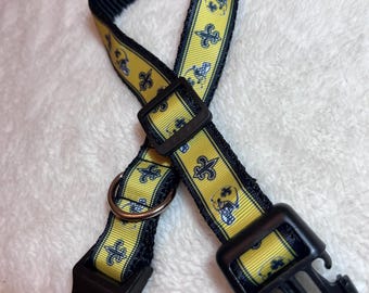 Könnte beinhalten: Ein schwarz-gelbes Hundehalsband mit einem Fleur-de-Lis-Muster. Das Halsband hat eine schwarze Kunststoffschnalle und einen Metallring zum Anbringen einer Leine. Das gelbe Band zeigt ein sich wiederholendes Muster aus blauen und weißen Fleur-de-Lis-Symbolen.