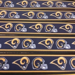 Könnte beinhalten: Ein marineblaues Ripsband mit einem sich wiederholenden Muster aus gelben und weißen Los Angeles Rams Football-Helm-Logos. Das Band hat dünne gelbe horizontale Streifen, die entlang der oberen und unteren Kanten des Stoffmaterials verlaufen.