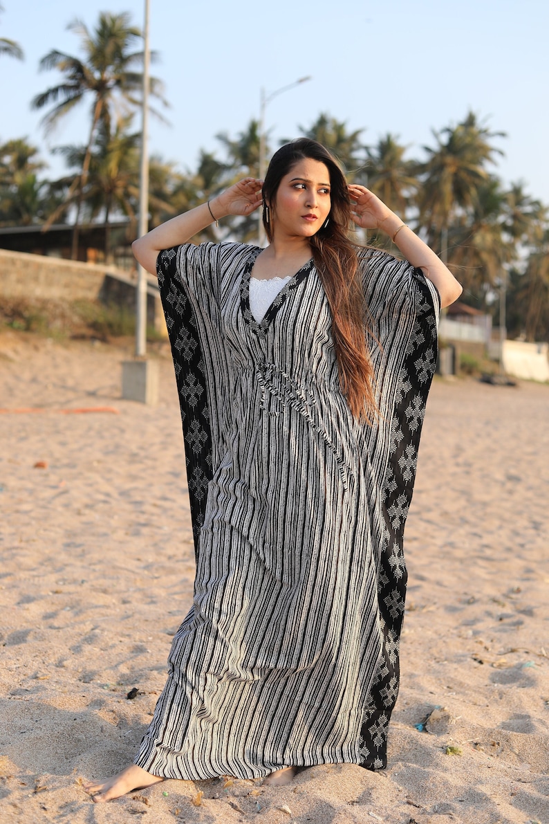 May include: A woman wearing a black and white striped kaftan with a V-neckline. The kaftan has a loose fit and is made of a lightweight fabric. The woman is standing on a sandy beach.