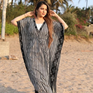 May include: A woman wearing a black and white striped kaftan with a white trim. The kaftan has a V-neck and is flowing. The woman is standing on a sandy beach.