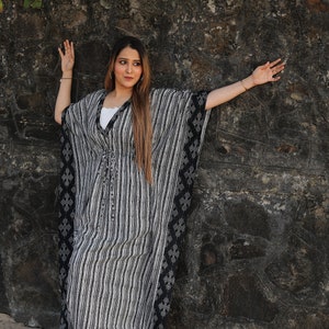 May include: A woman wearing a black and white striped kaftan with a black floral pattern on the sides. She is standing in front of a stone wall with green plants and pink flowers growing on top.