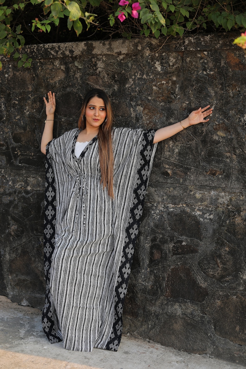 May include: A woman wearing a black and white striped kaftan with a black floral pattern on the sides. She is standing in front of a stone wall with green plants and pink flowers growing on top.