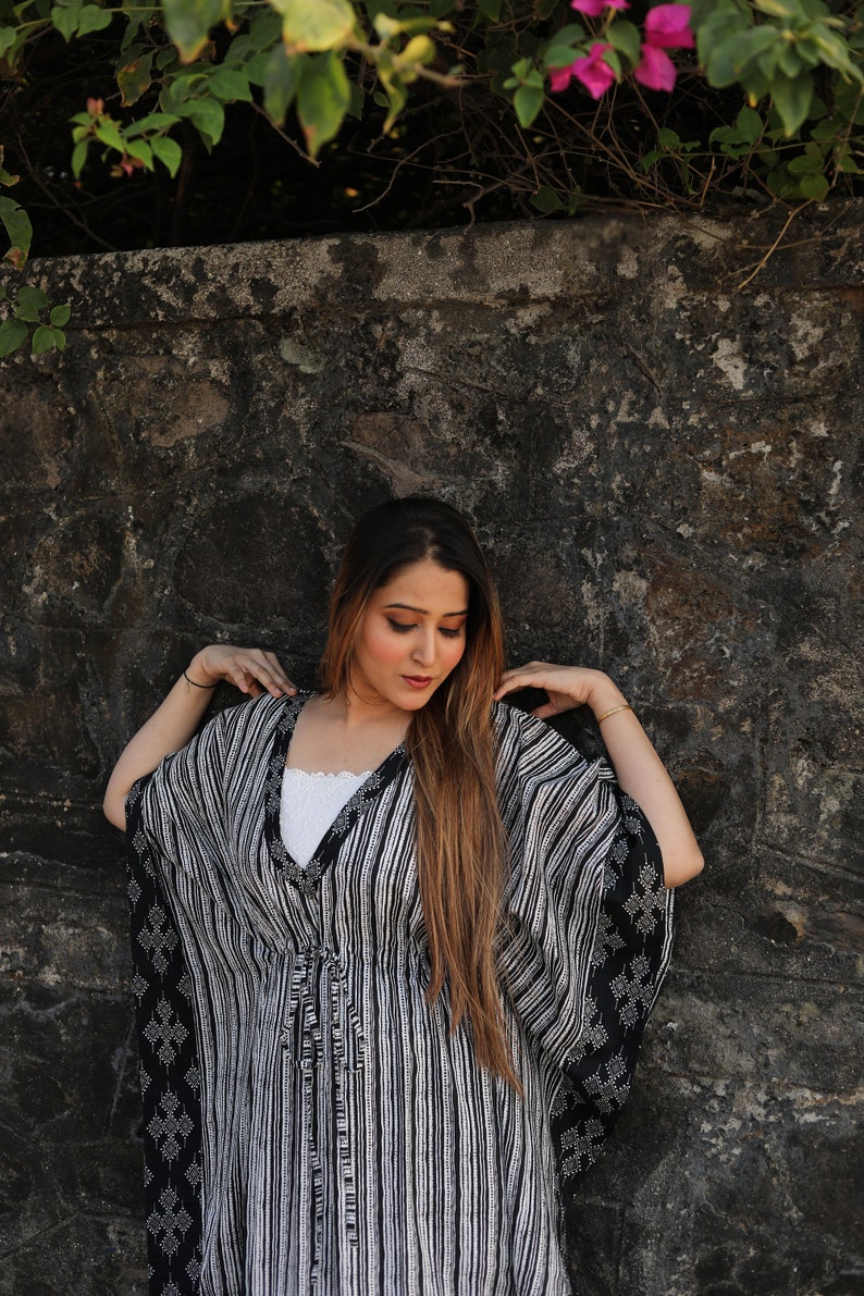 May include: A woman wearing a black and white striped kaftan with a white undergarment. The kaftan has a tie at the waist and a geometric pattern on the sleeves.