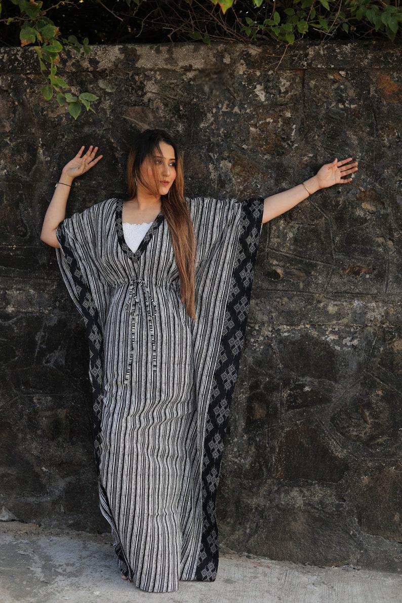 May include: A woman wearing a black and white striped kaftan with a black and white geometric pattern on the sleeves. The kaftan has a tie at the waist.