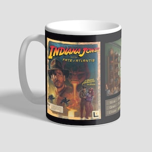 May include: A black and white mug with a graphic of Indiana Jones and the Fate of Atlantis video game cover. The mug has a white handle and a black interior.