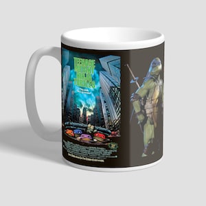 Pode incluir: Caneca de cerâmica branca com uma imagem preta e verde das Tartarugas Ninja. A imagem mostra as quatro tartarugas espiando por trás de um prédio e uma única tartaruga em pé em primeiro plano. O texto "Teenage Mutant Ninja Turtles" está impresso em verde na caneca.