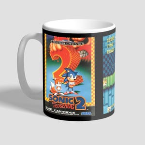 Könnte beinhalten: Weißer Keramikbecher mit dem Cover-Art des Videospiels Sonic the Hedgehog 2. Das Design zeigt den Spieletitel, die Charaktere Sonic und Tails sowie das Sega-Logo. Der Becher hat eine Standardgröße.