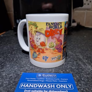 Könnte beinhalten: Eine weiße Keramiktasse mit einer bunten Illustration einer Comicfigur namens Dizzy aus dem Spiel "Fantasy World Dizzy". Die Tasse trägt den Text "Fantasy World Dizzy" und "94%". Die Tasse steht auf einer weißen Oberfläche mit einer blauen und weißen Karte, auf der "Nur Handwäsche (nicht für die Spülmaschine geeignet)" und "Etsy | Blueberryprintstore" steht.