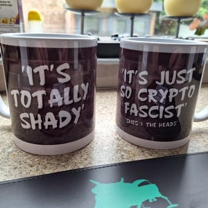 May include: Two white-handled coffee mugs with dark brown exteriors. One mug reads "IT'S TOTALLY SHADY" in white text. The other mug reads "IT'S JUST SO CRYPTO FASCIST" with "SMEG & THE HEADS" below.