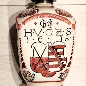 May include: A white ceramic bottle with a metal top, decorated with red and black designs. The bottle features a crest with the letters "HVCZS" and the number "1618". The design includes a shield and crossed swords.