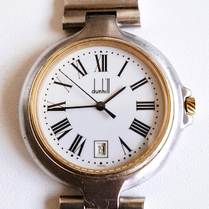 Könnte beinhalten: Eine Dunhill Armbanduhr mit rundem silberfarbenem Gehäuse, goldfarbener Lünette und weißem Zifferblatt mit schwarzen römischen Ziffern. Die Uhr verfügt über schwarze Zeiger für Stunden, Minuten und Sekunden, ein Datumsfenster und ein Metallgliederarmband.