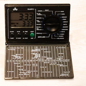 May include: Black quartz world time clock with a digital display showing 3:33 PM. Features buttons for clock, timer, and light, and a dial for time zones. Includes a world time zone map.