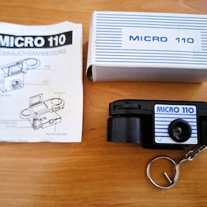 Pode incluir: Uma câmara miniatura preta Micro 110 com um porta-chaves, acompanhada pela sua caixa original às riscas brancas e azuis e um manual de instruções impresso com diagramas e o texto MICRO 110 GEBRAUCHSANWEISUNG.