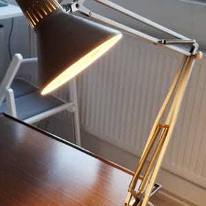 Könnte beinhalten: Eine graue Schreibtischlampe mit konischem Schirm und flexiblem Arm. Die Lampe strahlt helles Licht aus. Der Sockel ist an einem Holzschreibtisch befestigt. Das Design der Lampe erinnert an den Mid-Century-Modern-Stil.