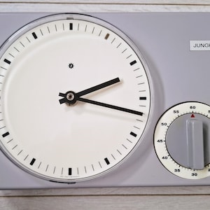 May include: A grey and white Junghans clock with a circular face and a separate timer dial. The clock face has black hands and markers, with the brand name "Junghans". The timer dial has a grey knob.