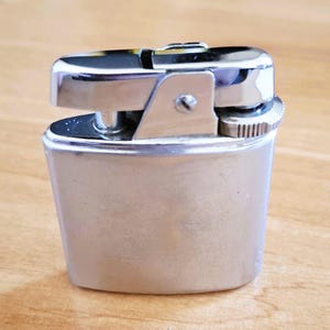 Può includere: Un accendino color argento con un corpo rettangolare e una parte superiore cromata. L'accendino ha un coperchio incernierato, una rotella di selce e un bocchettone per il carburante. Il design è compatto e sembra vintage.