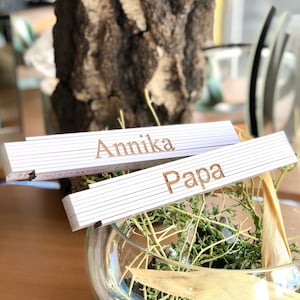 Könnte beinhalten: Zwei weiße, faltbare Holzlineale mit den eingravierten Namen "Annika" und "Papa". Die Lineale liegen in einer Glasvase mit getrockneten Pflanzen.