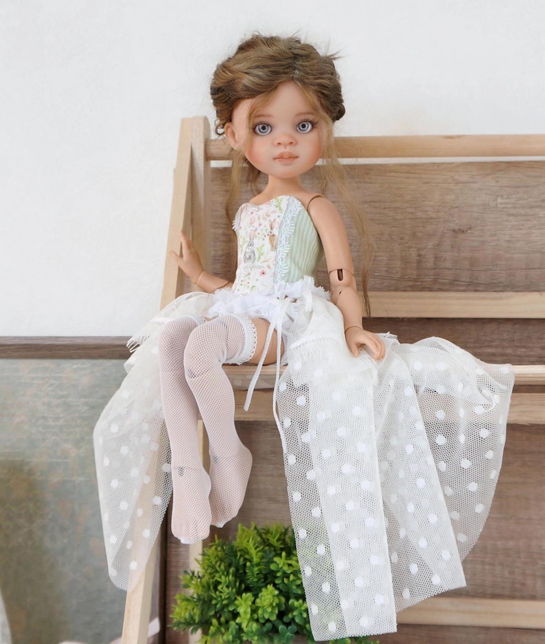 May include: A handcrafted doll with a detailed face, wearing a floral bodice, a white skirt with polka dots, and white thigh-high stockings. The doll has brown hair and is posed sitting on a wooden shelf. The doll's outfit is accented with lace.