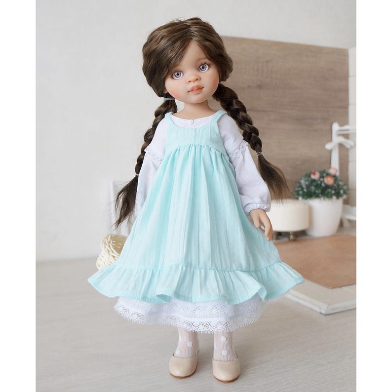 May include: A handcrafted doll with brown braided hair, blue eyes, and a serene expression. The doll wears a light blue dress over a white blouse and a white lace-trimmed skirt. Beige shoes complete the outfit. The doll is a charming collectible.