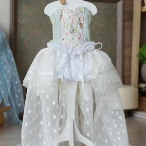 May include: A doll dress on a white stand. The dress features a bodice with a floral and bunny print, a ruffled neckline, and a layered skirt with white polka dots. A small heart-shaped tag with the text "It's My Heart" is attached.