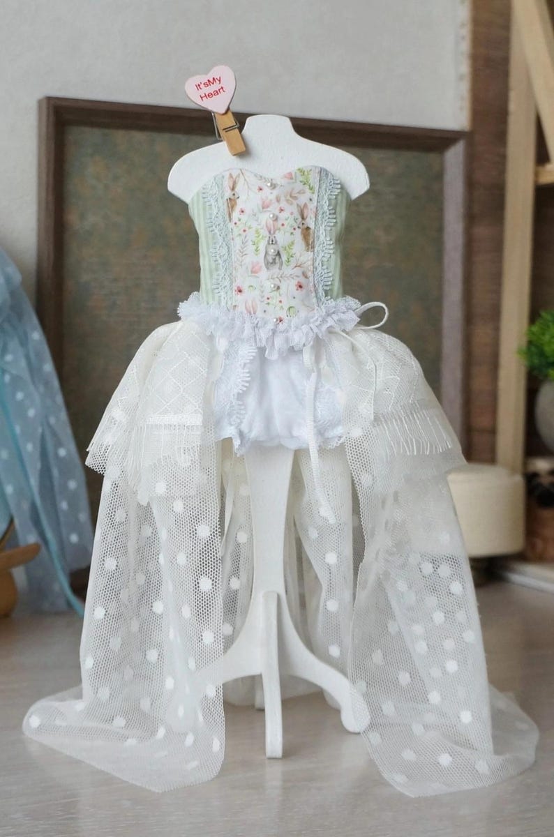 May include: A doll dress on a white stand. The dress features a bodice with a floral and bunny print, a ruffled neckline, and a layered skirt with white polka dots. A small heart-shaped tag with the text "It's My Heart" is attached.