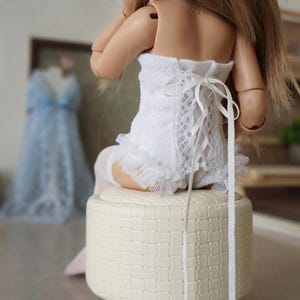 May include: A white lace corset with a ribbon tie-up back. The corset is part of a doll's outfit, paired with ruffled trim and white stockings. The doll is seated on a cream-colored, patterned stool.