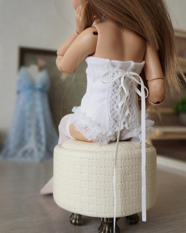 May include: A white lace corset with a ribbon tie-up back. The corset is part of a doll's outfit, paired with ruffled trim and white stockings. The doll is seated on a cream-colored, patterned stool.