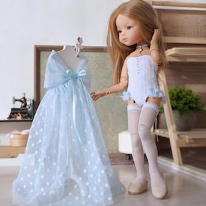 May include: A doll with long blonde hair stands next to a light blue dress with white polka dots. The dress is on a mannequin. The doll wears a white bodysuit, white stockings, and beige shoes. A small blue choker is around the doll's neck.