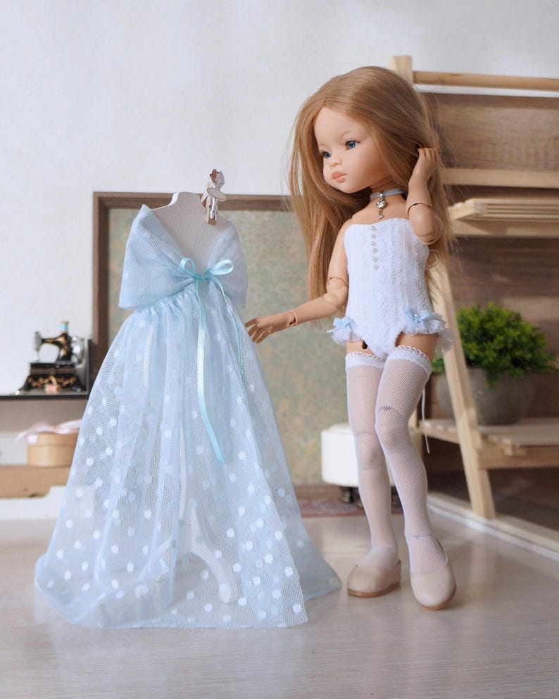 May include: A doll with long blonde hair stands next to a light blue dress with white polka dots. The dress is on a mannequin. The doll wears a white bodysuit, white stockings, and beige shoes. A small blue choker is around the doll's neck.