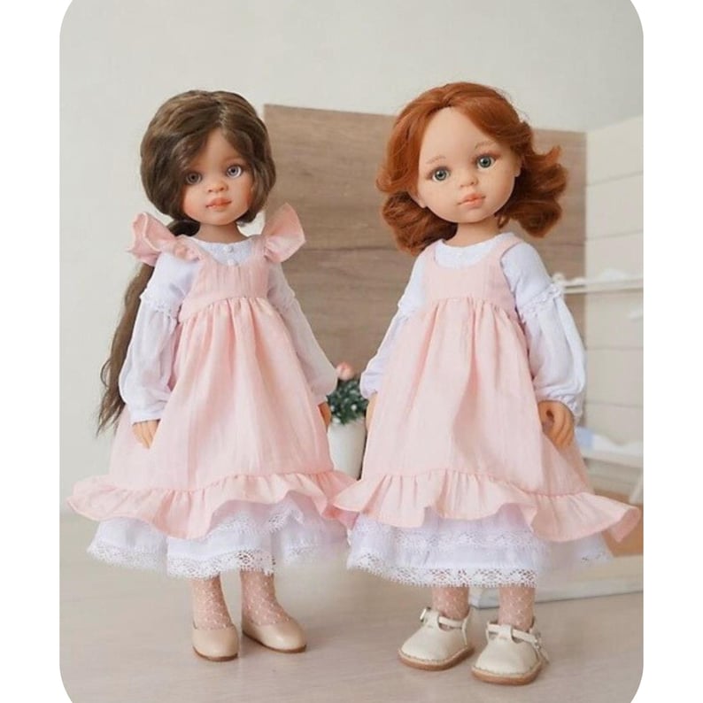 May include: Two dolls are displayed, each wearing a light pink dress with a white blouse underneath. The dresses have a ruffled hem and are paired with white shoes. One doll has brown hair, and the other has red hair.