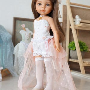 May include: A doll with brown hair styled in buns, wearing a white corset top with floral accents, white lace shorts, and a pink tulle skirt. The doll is also wearing white knee-high socks and beige shoes. A gold pendant is around the doll's neck.
