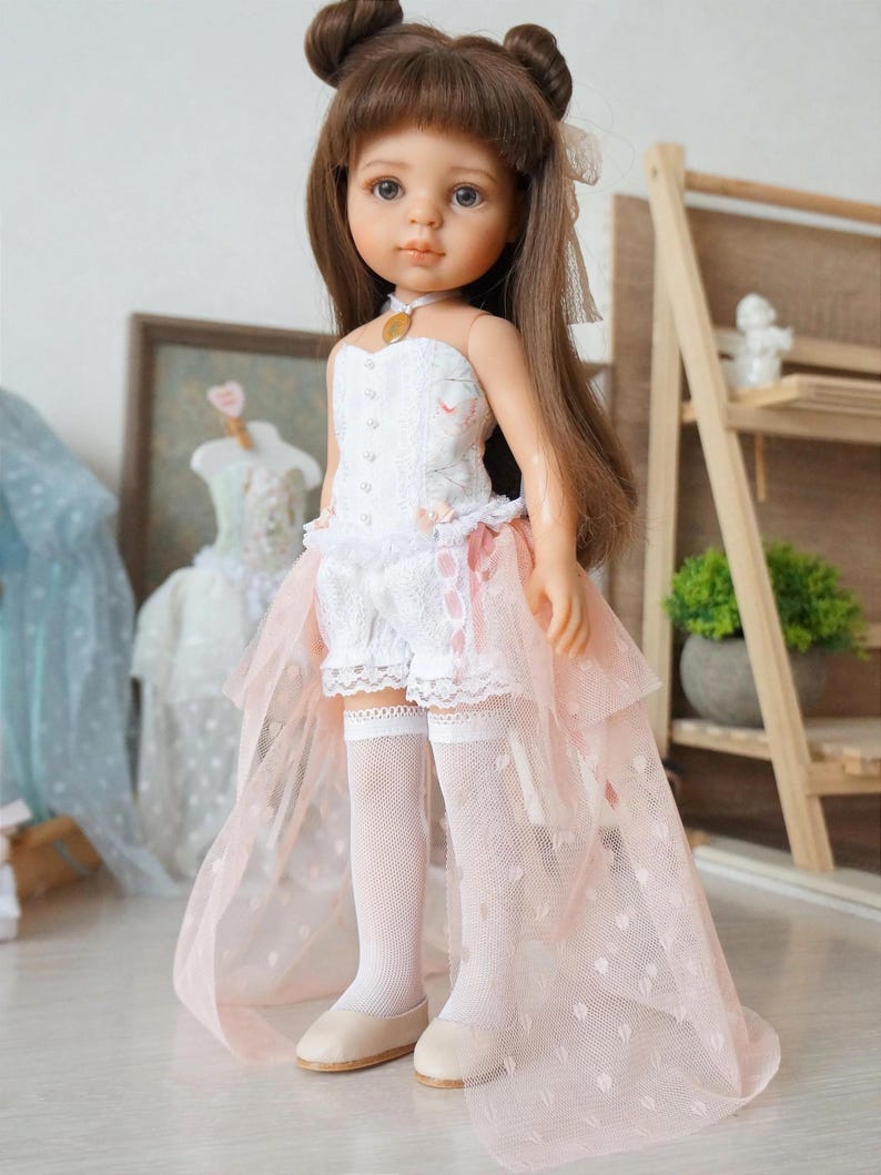 May include: A doll with brown hair styled in buns, wearing a white corset top with floral accents, white lace shorts, and a pink tulle skirt. The doll is also wearing white knee-high socks and beige shoes. A gold pendant is around the doll's neck.