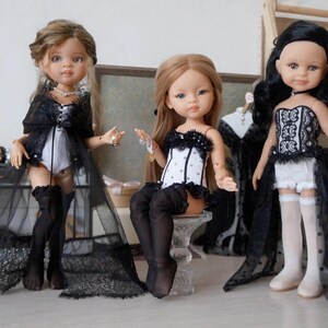 May include: Three fashion dolls are displayed, each with unique outfits. The dolls wear black and white lingerie-style ensembles with lace accents. One doll has a long, sheer black overcoat. The dolls have different hairstyles and accessories.