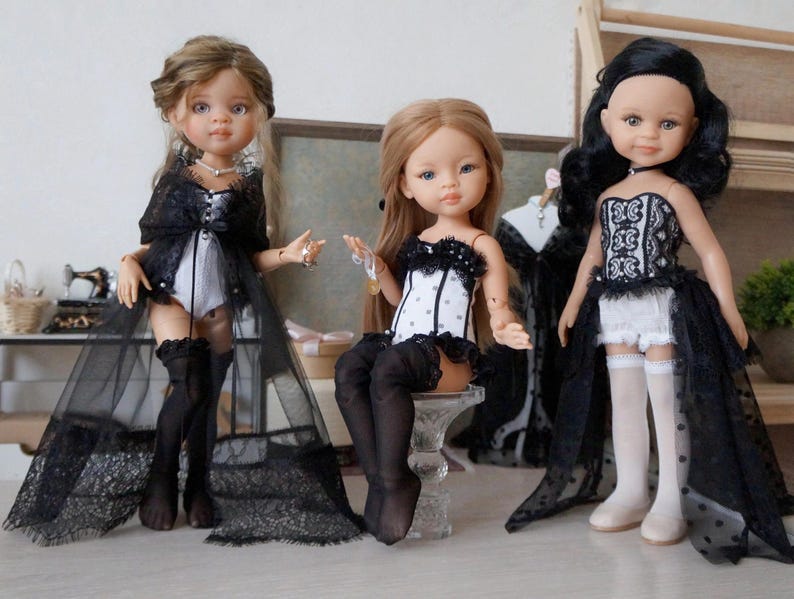 May include: Three fashion dolls are displayed, each with unique outfits. The dolls wear black and white lingerie-style ensembles with lace accents. One doll has a long, sheer black overcoat. The dolls have different hairstyles and accessories.