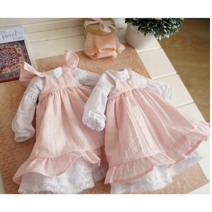 May include: Two pink dresses with white long sleeves and lace trim. The dresses have a ruffled skirt and a bow detail on the shoulder. The dresses are displayed on a light-colored surface.
