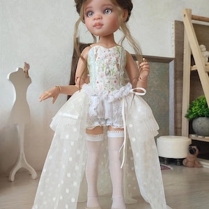 May include: A doll with light brown hair and blue eyes, wearing a white dress with a floral bodice and a long, sheer, polka-dot train. The doll is also wearing white shorts, stockings, and beige shoes. The doll is standing in front of a light-colored wall.