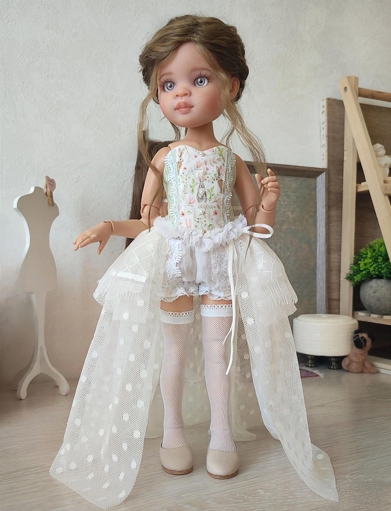 May include: A doll with light brown hair and blue eyes, wearing a white dress with a floral bodice and a long, sheer, polka-dot train. The doll is also wearing white shorts, stockings, and beige shoes. The doll is standing in front of a light-colored wall.