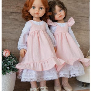 May include: Two dolls with realistic features stand side-by-side. They have detailed faces, with one doll having red hair and the other brown. Both dolls wear pink dresses with white lace trim and white shoes. The dolls are a charming addition to any collection.