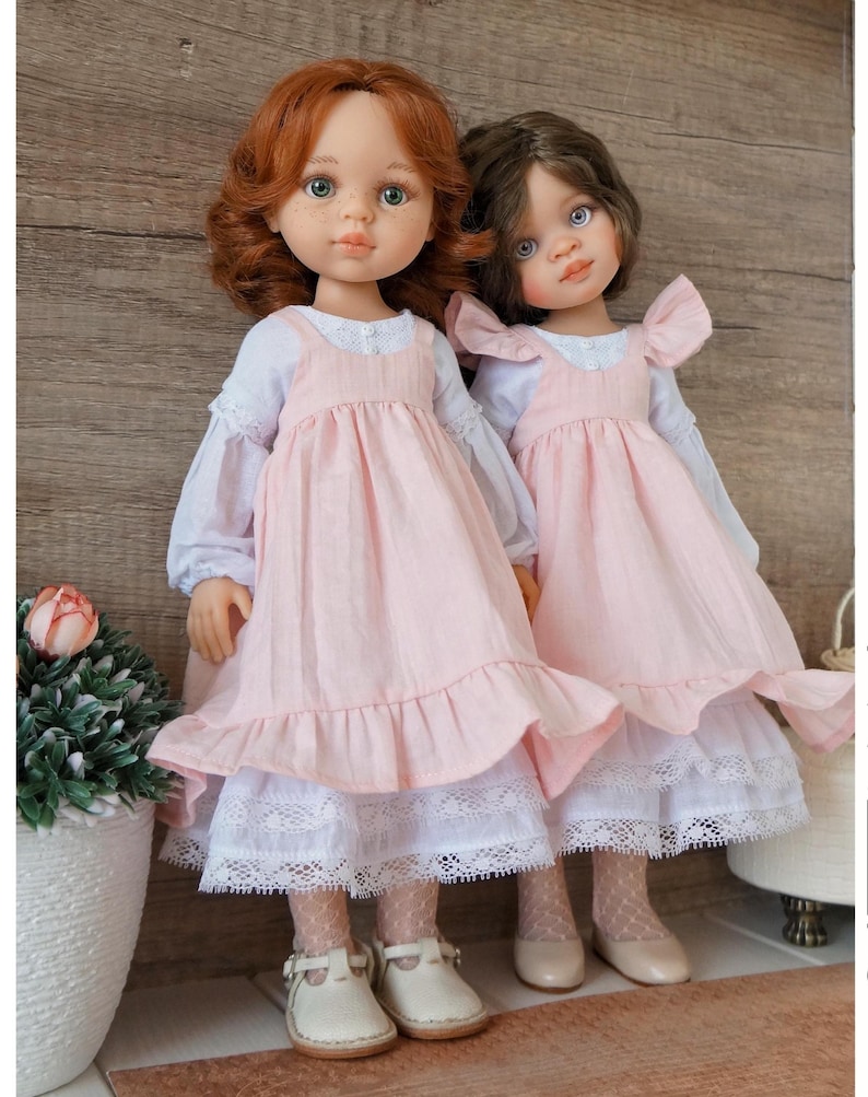 May include: Two dolls with realistic features stand side-by-side. They have detailed faces, with one doll having red hair and the other brown. Both dolls wear pink dresses with white lace trim and white shoes. The dolls are a charming addition to any collection.