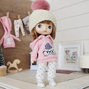 Puede incluir: Una muñeca con una sudadera rosa con un diseño de cara de gato, pantalones blancos con lunares y zapatillas blancas. La muñeca lleva un gorro de punto color crema con un pompón rosa. Ropa de muñeca adicional cuelga en el fondo.