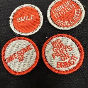 Peut inclure: Quatre patchs brodés avec des coutures rouges et des lettres blanches. Les patchs sont sur un fond de tissu noir. Les patchs indiquent "Smile", "Chin Up Tits Out It's All Good", "Awesome AF", et "Big Girl Pants On Bring It".