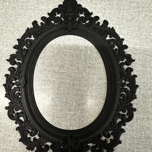 May include: Black oval picture frame with intricate, swirling patterns. The frame has a matte finish and a vintage style. The frame is empty, ready to display a photograph or artwork.