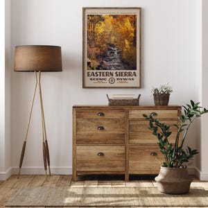May include: A framed vertical wall art print titled Eastern Sierra Scenic Byway featuring a photograph of a flowing stream surrounded by golden autumn trees. The poster includes text detailing the location and establishment date of the route.