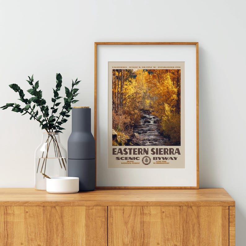 May include: A framed vertical poster of the Eastern Sierra Scenic Byway featuring a photograph of a stream surrounded by golden autumn trees. The text reads Eastern Sierra Scenic Byway, Inyo National Forest, Lone Pine to Bridgeport.