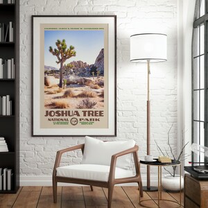 May include: Framed poster featuring a landscape of Joshua Tree National Park. The poster has a beige border and includes the park's name and logo. The image shows a Joshua tree, rocks, and desert vegetation under a blue sky. The poster is hanging on a white brick wall.