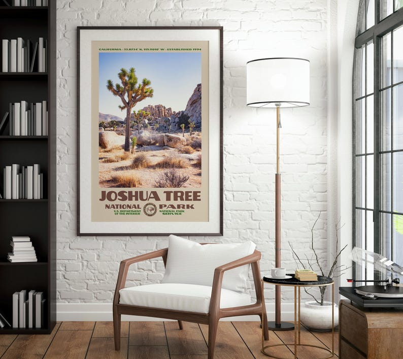 May include: Framed poster featuring a landscape of Joshua Tree National Park. The poster has a beige border and includes the park's name and logo. The image shows a Joshua tree, rocks, and desert vegetation under a blue sky. The poster is hanging on a white brick wall.