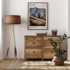 May include: A framed print of Half Dome in Yosemite National Park hangs above a wooden dresser. The print features a landscape photograph with the text "HALF DOME" at the top. A floor lamp with a brown shade and a woven basket are also visible.